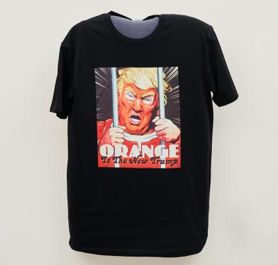 A black t-shirt with an image of an orange-faced man wearing orange and looking angry, both hands clinging to metal bars in front of him, with the words Orange Is The New Trump.